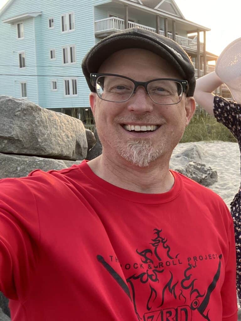 AW Galuszka smiling in a red shirt wearing eyeglasses and a flat cap. in Carolina Beach NC with a sea side blue condo in the background.